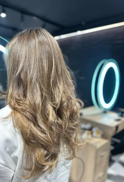 Balayage sur longueurs ondulées, devant un miroir rond cerclé de lumière turquoise du salon Gem Coiffure à Borgo